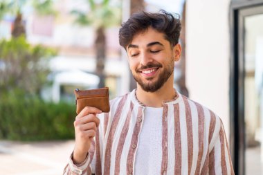 Yakışıklı bir Arap açık havada mutlu bir ifadeyle cüzdan tutuyor.