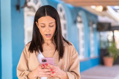 Dışarıda kameraya bakan genç bir kadın şaşırmış bir ifadeyle cep telefonunu kullanıyor.