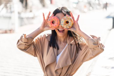 Açık havada genç bir kadın gözlerinde donut tutuyor.