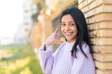 Açık havada genç bir kadın telefon jesti yapıyor. Bana geri dönüş işareti ver.