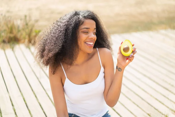 stock image Young African American woman holding an avocado at outdoors with happy expression