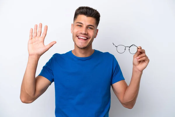 白い背景に隔離された眼鏡を持つ若い白人男性幸せな表情で手で敬礼 — ストック写真