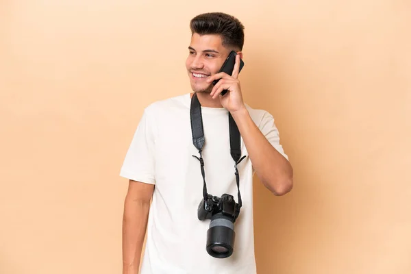 年轻的摄影师在米色背景下被隔离的高加索人与他人在手机上交谈 — 图库照片