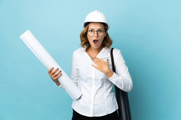 Young architect Georgian woman with helmet and holding blueprints over isolated background surprised and shocked while looking right