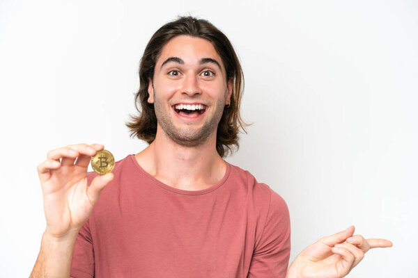 Молодой красивый мужчина, держащий биткоин на белом фоне, удивлённый и указывающий пальцем в сторону