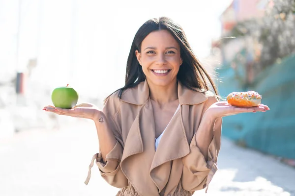 Young woman at outdoors holding apple and donut with happy expression