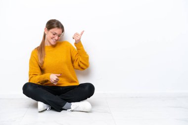 Beyaz arka planda, yerde oturan beyaz tenli genç bir kadın gitar hareketi yapıyor.