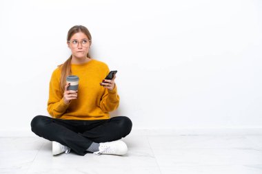 Beyaz arka planda izole bir şekilde yerde oturan beyaz tenli genç bir kadın elinde kahve ve bir şeyler düşünürken bir cep telefonu tutuyordu.