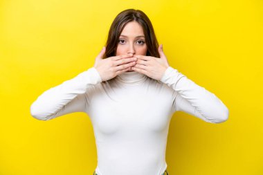 Sarı arka planda izole edilmiş genç beyaz kadın elleriyle ağzını kapatıyor.