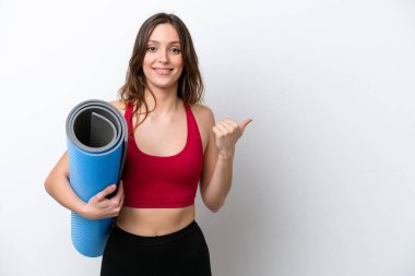 Young sport caucasian woman going to yoga classes while holding a mat isolated on white background pointing to the side to present a product