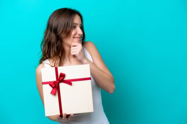 Kafkasyalı genç bir kadın mavi arka planda izole edilmiş bir hediye tutuyor. Bir fikir üretiyor ve bakıyor.