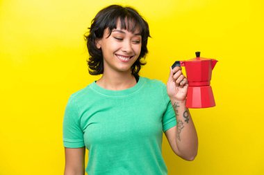 Arjantinli genç bir kadın elinde kahve makinesini tutarken mutlu bir ifadeyle sarı arka planda izole edilmiş.