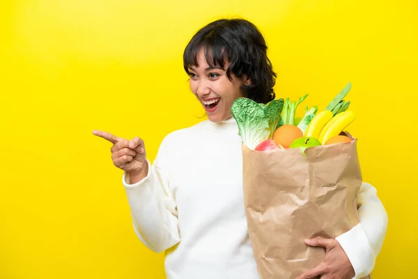 年轻的阿根廷女人拿着一个用黄色背景隔开的购物袋 手指指向旁边 并展示了一种产品 — 图库照片