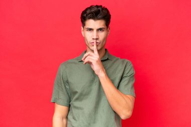 Kırmızı arka planda izole edilmiş beyaz, yakışıklı genç bir adam sessizlik işareti gösteriyor. Parmağını ağzına sokuyor.