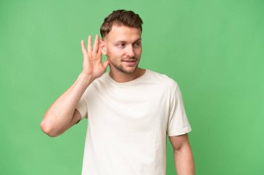 Sarışın, beyaz bir adam izole edilmiş arka planda bir şeyler dinlerken elini kulağına koyar.