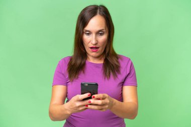 Orta yaşlı beyaz kadın izole edilmiş arka planda kameraya bakarken şaşırmış bir ifadeyle cep telefonuna bakıyor.