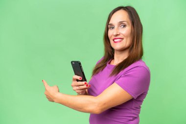 Orta yaşlı beyaz kadın izole edilmiş arka planda cep telefonu kullanıyor ve geriye doğru işaret ediyor.