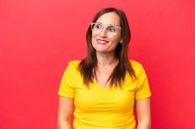 Orta yaşlı beyaz kadın, kırmızı arka planda izole edilmiş, yana bakıyor ve gülümsüyor.