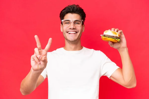 Młody Biały Mężczyzna Trzyma Hamburgera Odizolowany Czerwonym Tle Uśmiechając Się — Zdjęcie stockowe