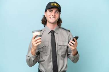 Genç güvenlik görevlisi mavi arka planda izole edilmiş. Elinde kahve ve bir şeyler düşünürken cep telefonu var.