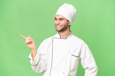 Genç, yakışıklı aşçı, izole edilmiş arka planda. Parmakla göstererek bir ürün sunuyor.