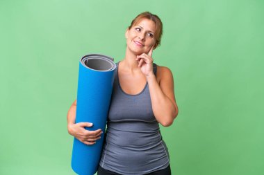 Orta yaşlı sporcu bir kadın yoga derslerine giderken elinde paspasla izole bir arka planda bir fikir ararken kafasını kaldırıp yukarı bakıyor.