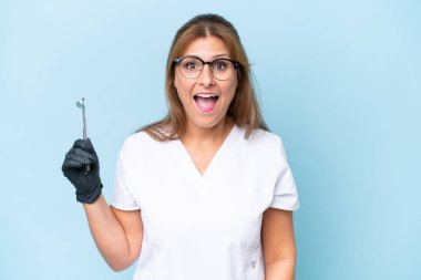 Orta yaşlı Dişçi, mavi arka plan üzerinde sürpriz yüz ifadesi ile araçlar tutuyor