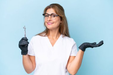 Orta yaşlı dişçi mavi arka plan üzerinde araçlar tutuyor. Davet etmek için ellerini yana uzatıyor.