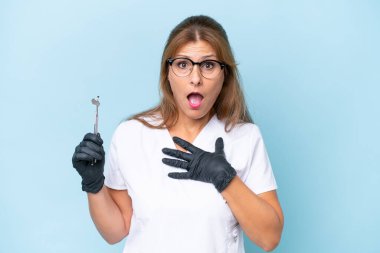 Orta yaşlı dişçi mavi arka plan üzerinde araçlar tutuyor sağa bakarken şaşırmış ve şok olmuş.