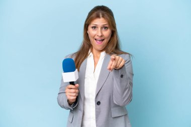 İzole edilmiş mavi arka planda orta yaşlı televizyon sunucusu kadın şaşırmış ve ön tarafı işaret ediyor.