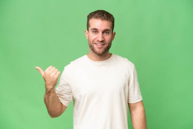 Yeşil krom arka planda izole edilmiş yakışıklı beyaz adam bir ürün sunmak için kenarı işaret ediyor.