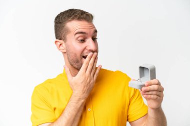 Young man holding a engagement ring isolated on white background with surprise and shocked facial expression