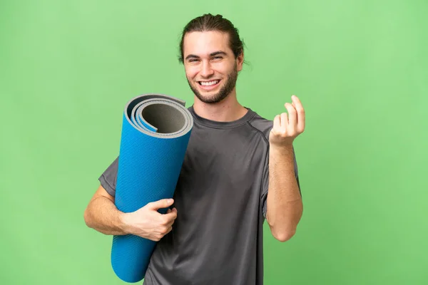 年轻的运动员一边去上瑜伽课 一边拿着垫子挡住孤立的背景 做着赚钱的手势 — 图库照片