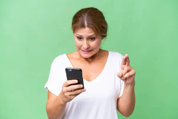 stock image Middle-aged caucasian woman over isolated background using mobile phone with fingers crossing