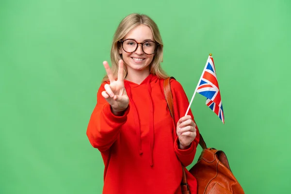 孤立した背景に英国の旗を掲げる若いヒスパニック系女性が笑顔と勝利の兆候を示す — ストック写真