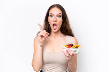 Beyaz arka planda bir kase meyve tutan genç beyaz kadın çözümün farkına varmak için parmağını kaldırıyor.