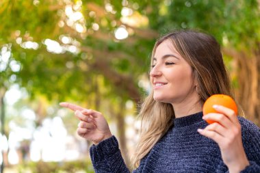 Genç, güzel Romen bir kadın elinde portakal tutarak bir ürünü sunmak için kenarı işaret ediyor