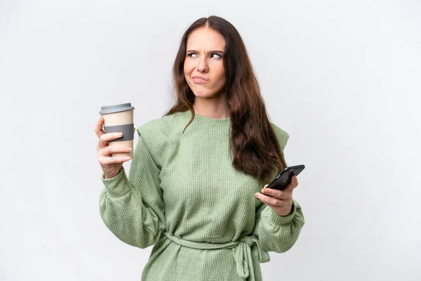 年轻的高加索女人被隔离在白色背景下 一边拿着咖啡 一边想着什么 一边拿着手机 — 图库照片