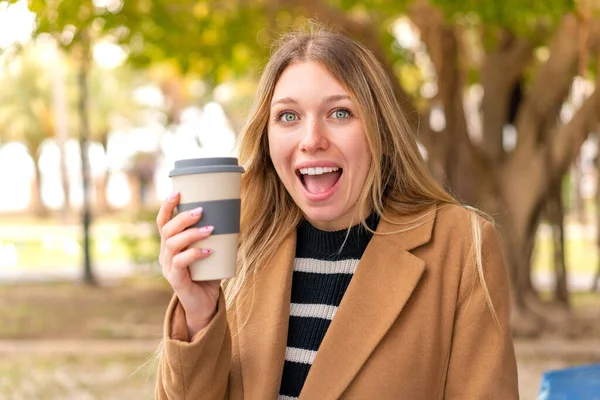 年轻漂亮的金发女子在户外端着咖啡 脸上带着惊讶和震惊的表情 — 图库照片