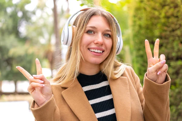Νεαρή Όμορφη Ξανθιά Γυναίκα Ακούει Μουσική Ακουστικά Εξωτερικούς Χώρους Δείχνει — Φωτογραφία Αρχείου