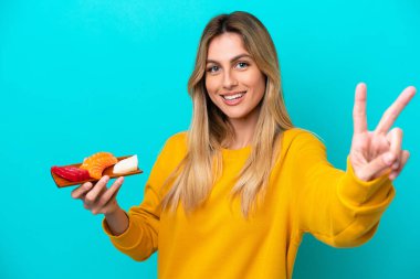 Mavi arka planda saşimi tutan genç Uruguaylı kadın gülümsüyor ve zafer işareti gösteriyor.