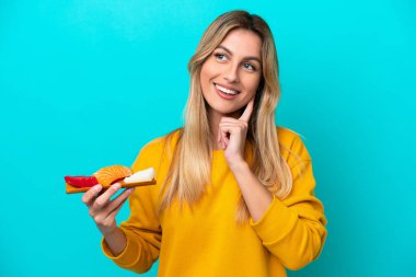 Genç Uruguaylı kadın elinde saşimi tutarken mavi arka planda izole edilmiş bir şekilde yukarı bakarken bir fikir düşünüyor.