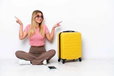 Uruguaylı gezgin kadın, beyaz arka planda izole bir şekilde yerde oturuyor. Yan tarafları işaret ediyor ve mutlu.