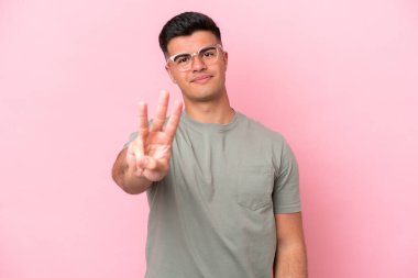 Beyaz tenli yakışıklı adam pembe arka planda izole edilmiş mutlu ve parmaklarıyla üçe kadar sayıyor.