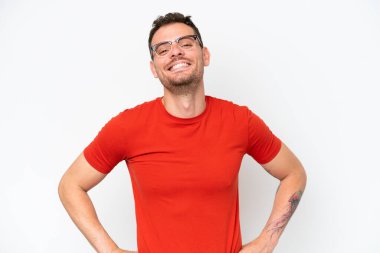 Beyaz arka planda soyutlanmış beyaz tenli yakışıklı genç adam kolları kalçasında ve gülümsüyor.