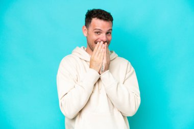 Genç, beyaz, yakışıklı bir adam mavi arka planda izole edilmiş mutlu ve gülümseyen elleriyle ağzını kapatıyor.