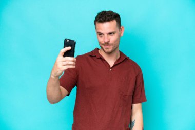 Mavi arka planda izole edilmiş beyaz, yakışıklı genç bir adam selfie çekiyor.