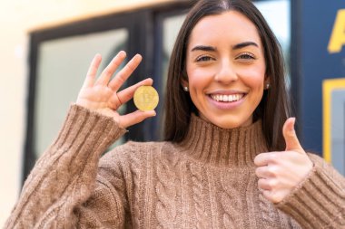 Dışarıda Bitcoin tutan genç bir kadın. Başparmakları havada. Çünkü iyi bir şey oldu.