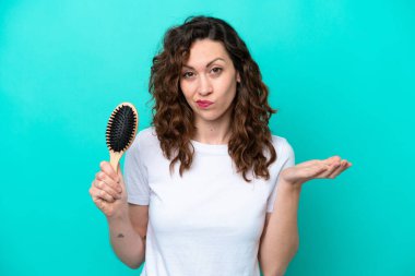 Mavi arka planda saç fırçası tutan beyaz bir kadın omuzlarını kaldırırken şüpheyle hareket ediyor.