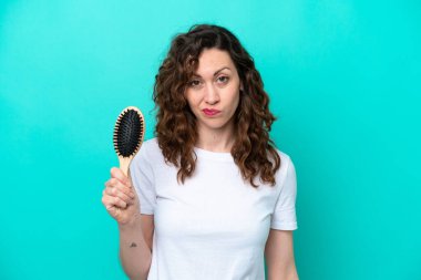 Mavi arka planda saç fırçası tutan beyaz bir kadın hüzünlü bir ifadeyle izole edilmiş.